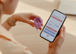Woman holding a menstrual cup as she checks her cycle calendar app on the phone.